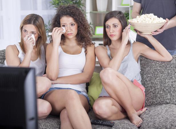 Drei junge Frauen sitzen auf einem Sofa und weinen, während ein Mann Popcorn bringt.