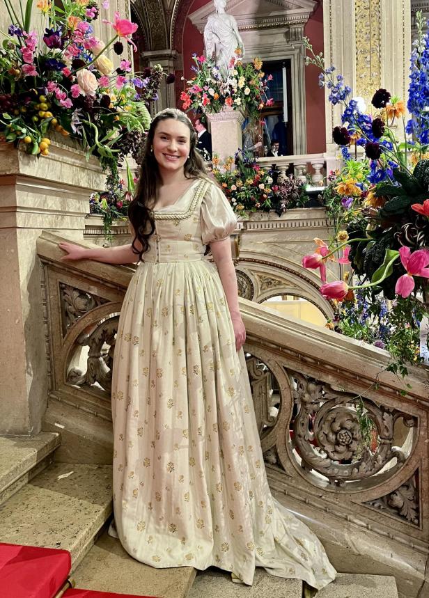 Eine junge Frau in einem historischen Kleid steht auf einer verzierten Treppe, umgeben von Blumenarrangements.