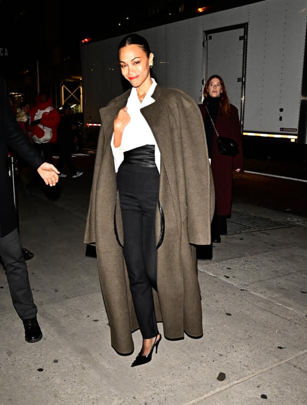 Zoe Saldana trägt einen langen Mantel über einem weißen Hemd und einer schwarzen Hose.