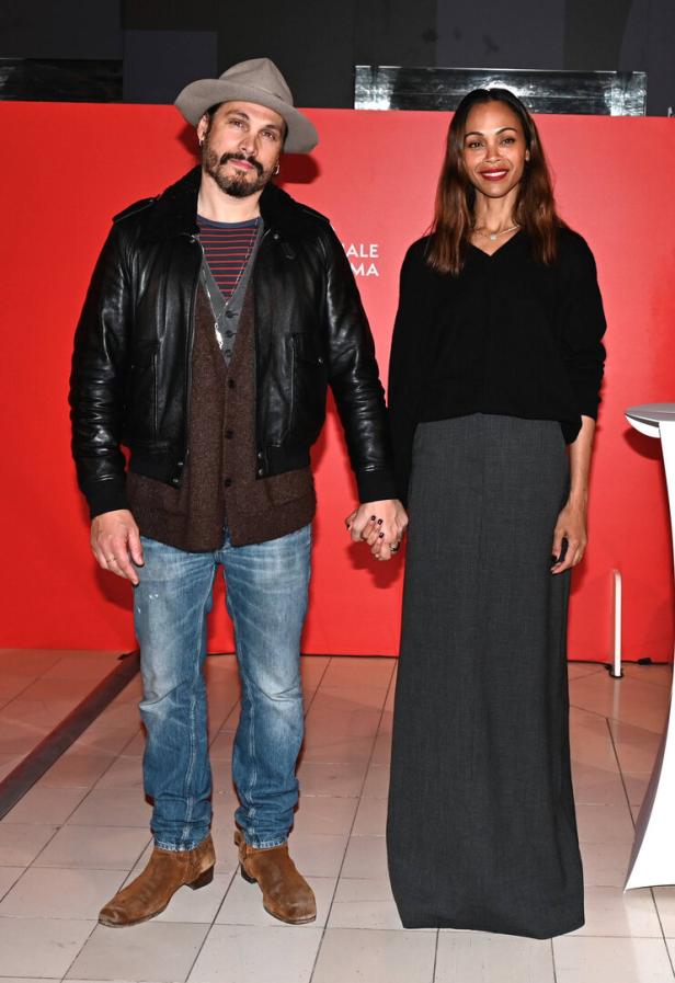 Marco Perego und Zoe Saldana halten Händchen vor einem roten Hintergrund.