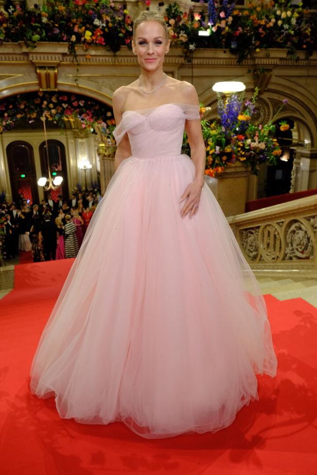 Eine Frau in einem rosa Ballkleid steht auf einem roten Teppich vor einer festlichen Kulisse.