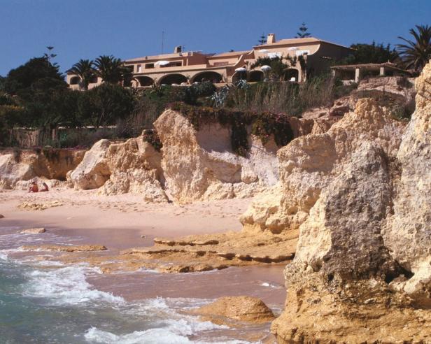 Ein Strand mit Sandsteinfelsen und einem Gebäude auf einer Klippe im Hintergrund.