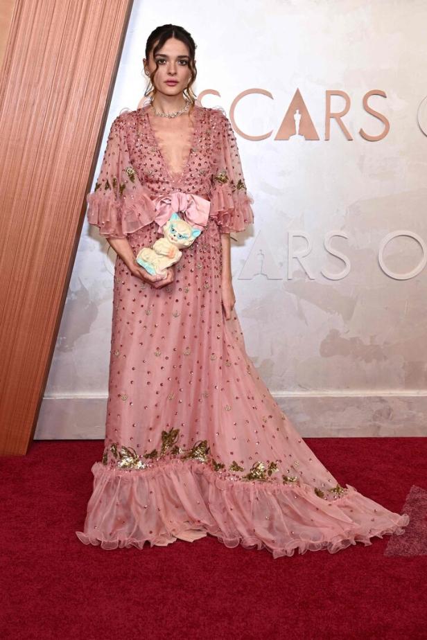 Eine Frau in einem rosa Kleid hält eine Katzenfigur auf dem roten Teppich der Oscars.