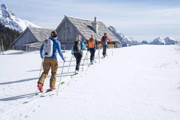 Vier Personen gehen auf Skiern in einer verschneiten Landschaft an Holzhütten vorbei.
