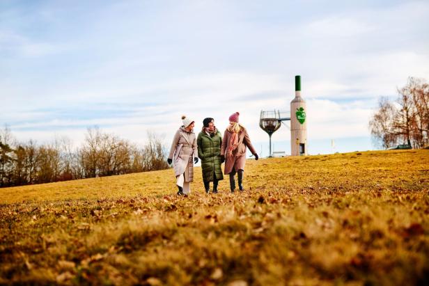 Drei Frauen spazieren auf einem Feld mit einer großen Weinflaschen-Skulptur im Hintergrund.