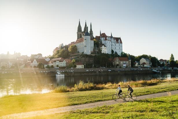 Zwei Fahrradfahrer fahren an einem Fluss entlang. Hinter ihnen ragt eine Burg empor.