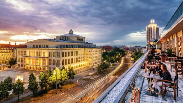 Eine Hauptstraße von einer Dachterrasse aus gesehen, hinter der Hauptstraße wird im Abendlicht eine Oper bestrahlt.