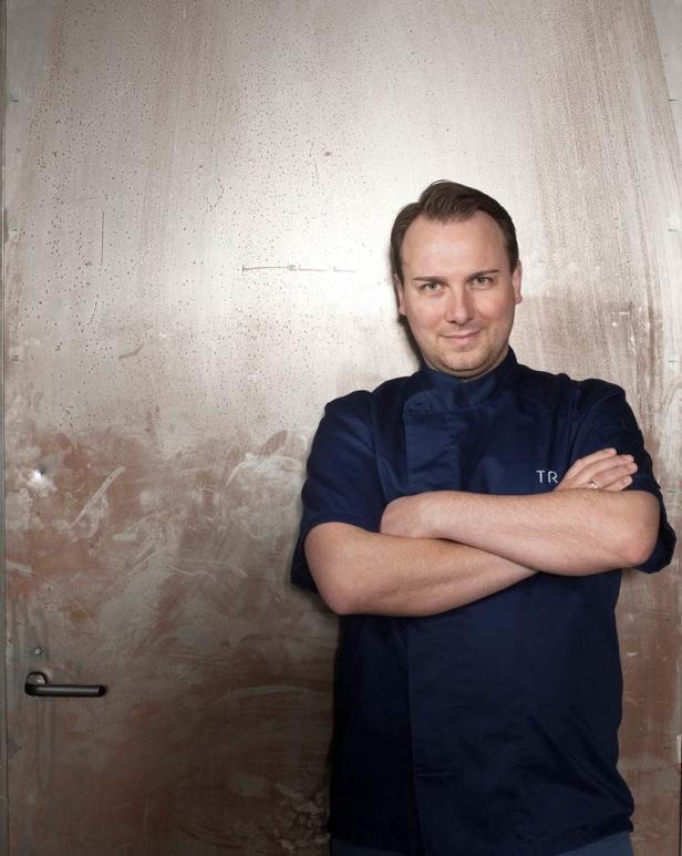 Ein Mann in Kochuniform steht vor einer Tür und hat die Arme verschränkt.