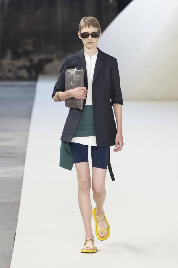 Eine Person mit Sonnenbrille läuft in einem Blazer und gelben Sandalen über einen Laufsteg.