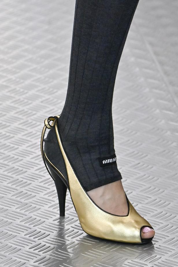 Ein Fuß in einer grauen Leggings und einem goldenen Miu Miu-Pumps mit Peeptoe.