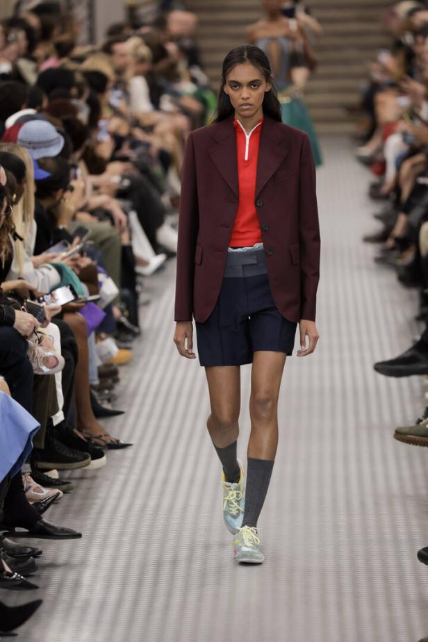 Eine Frau präsentiert ein Outfit mit Blazer, Shorts, Socken und Turnschuhen auf einem Laufsteg.