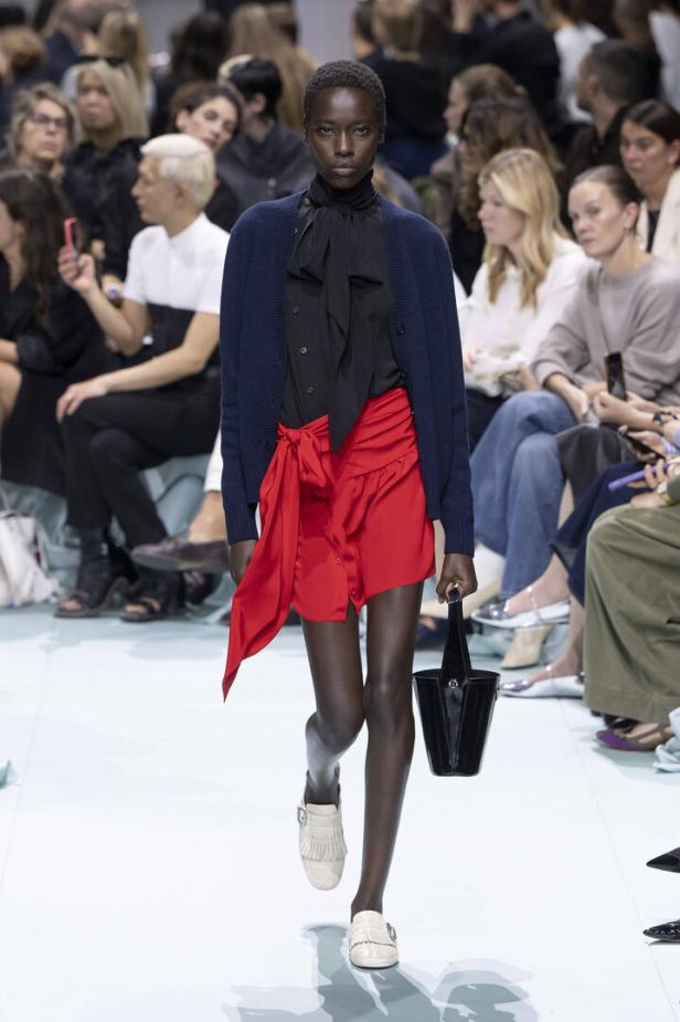 Eine Frau in einem roten Rock, einer schwarzen Bluse und einer marineblauen Strickjacke auf einem Laufsteg.