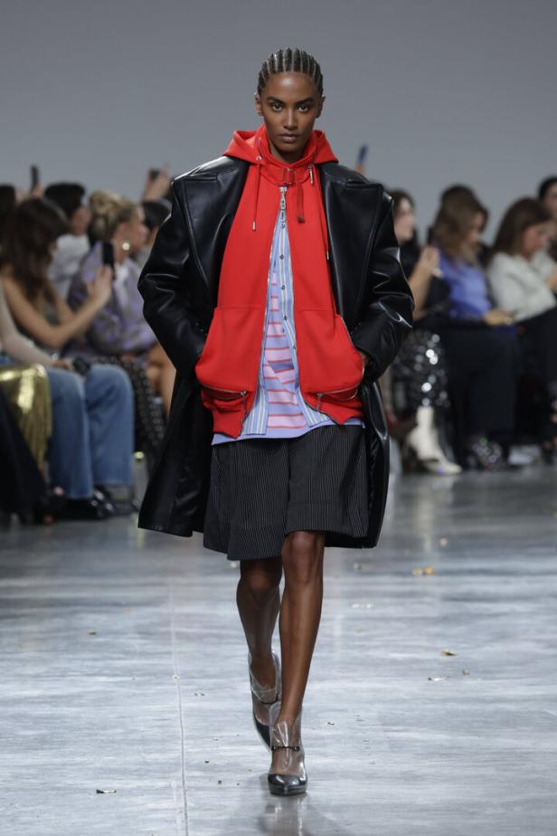 Ein Model auf dem Laufsteg trägt einen roten Hoodie, einen schwarzen Ledermantel und transparente Schuhe.