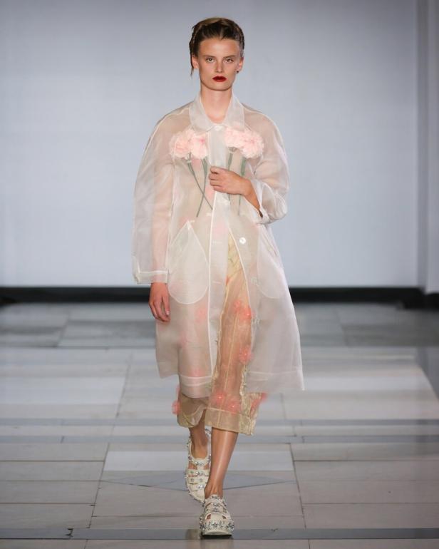 Ein Model auf dem Laufsteg trägt einen transparenten Mantel mit Blumenapplikationen und hält rosa Blumen.
