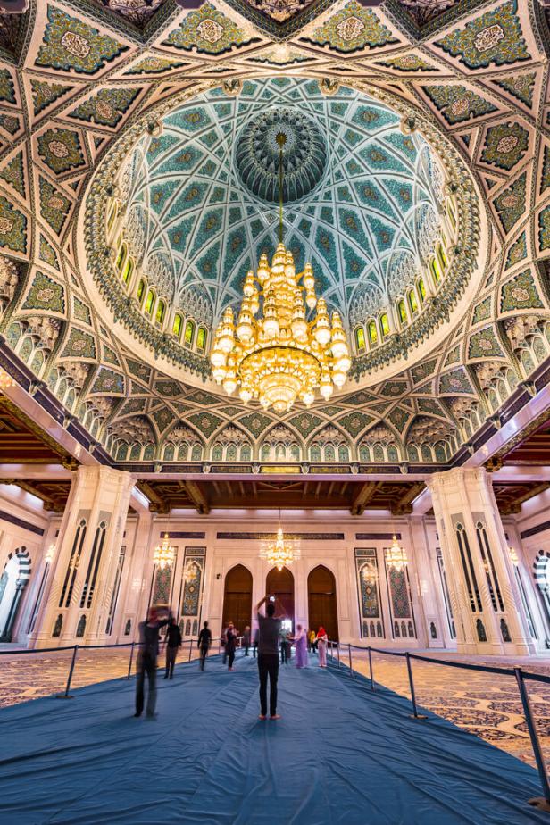 Blick in die prunkvolle Sultan-Qabus-Moschee mit Menschen unter der verzierten Kuppel und dem Kronleuchter.