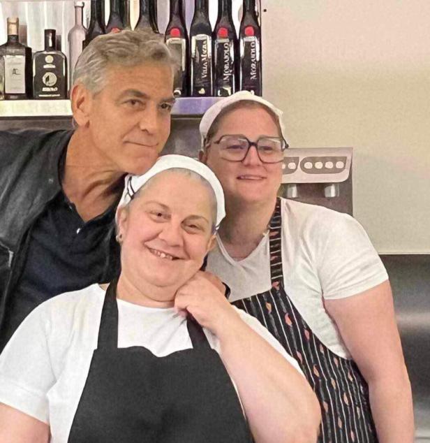 George Clooney posiert mit zwei Frauen in Kochkleidung vor einer Bar.