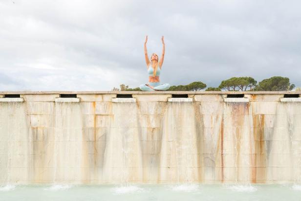 Eine Frau meditiert mit erhobenen Armen auf einer Mauer mit Wasserfällen.