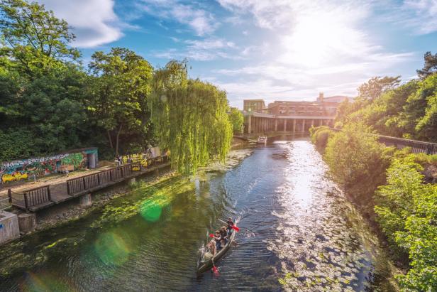 Ein Kanal, auf dem Ruderer sind. Daneben Bäume. Im Hintergrund Häuser.