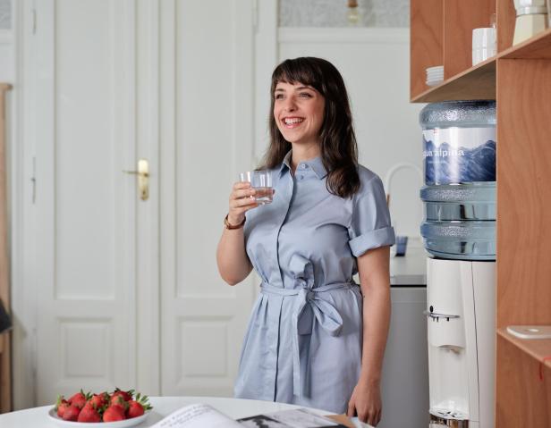 Eine Frau steht neben einem Wasserspender und lächelt, während sie ihr Glas Wasser in der Hand hält.