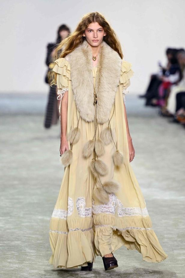 Ein Model präsentiert ein langes, hellgelbes Kleid mit Fellkragen auf dem Laufsteg.