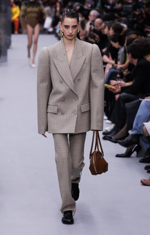 Ein Model präsentiert einen übergroßen Hosenanzug mit Hahnentrittmuster und eine braune Handtasche auf dem Laufsteg.