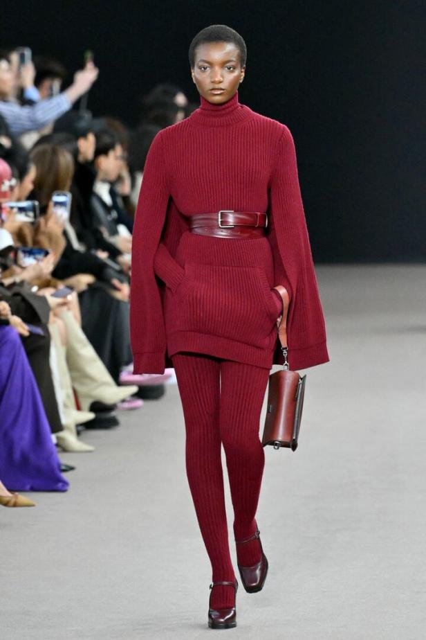 Eine Frau in einem roten Strickensemble mit Gürtel und Handtasche auf einem Laufsteg.
