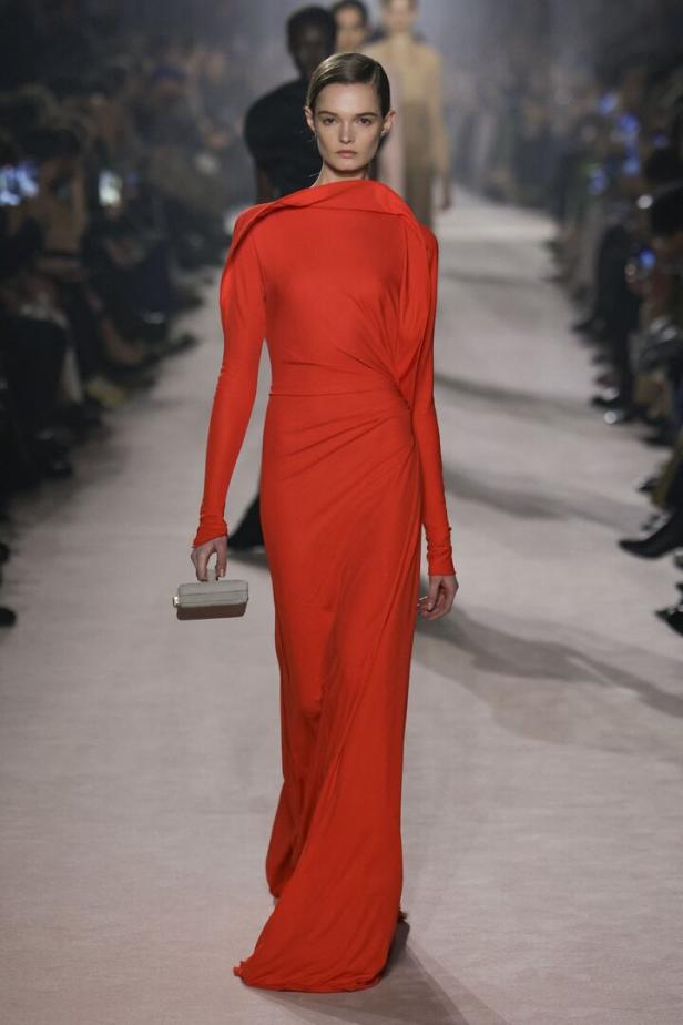 Ein Model präsentiert ein langes, rotes Kleid mit Drapierung auf dem Laufsteg.
