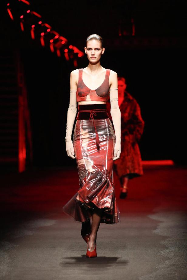Ein Model präsentiert ein rotes Outfit mit transparentem Rock auf einem Laufsteg.