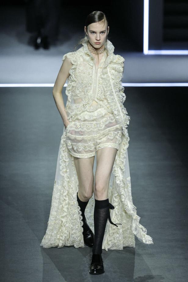 Ein Model präsentiert ein weißes, aufwendig verziertes Spitzenensemble mit Rüschen und schwarzen Kniestrümpfen auf dem Laufsteg.