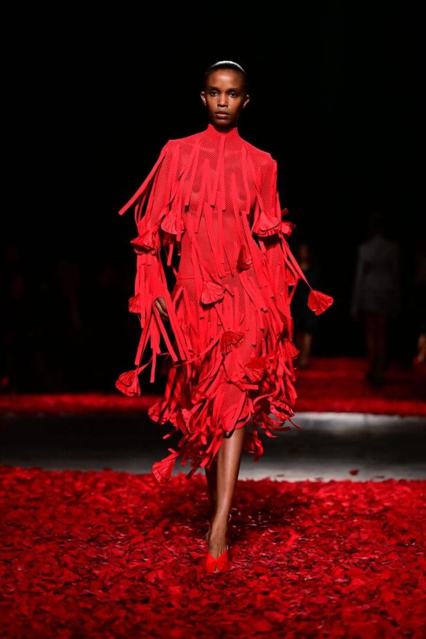 Eine Frau in einem roten Fransenkleid auf einem Laufsteg voller roter Blütenblätter.