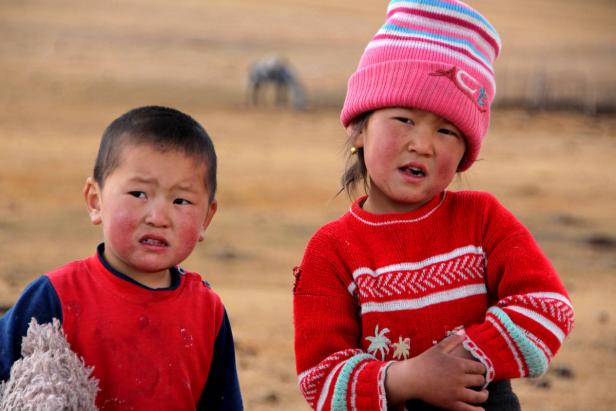 Zwei Kinder stehen in einer kargen Landschaft, ein Pferd ist im Hintergrund zu sehen.