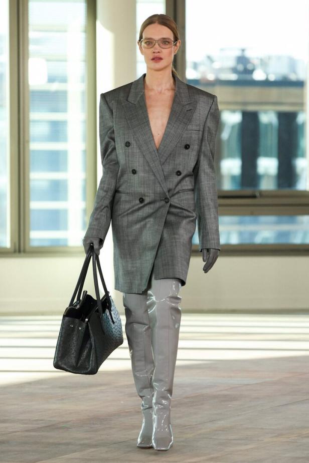 Eine Frau mit Brille trägt einen übergroßen Blazer, Overknee-Stiefel und eine Handtasche.