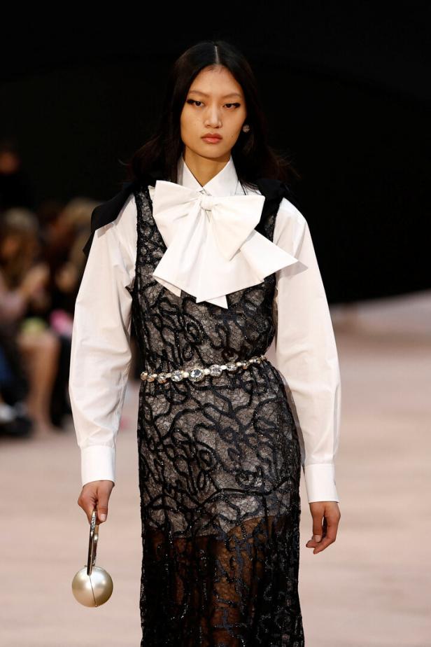 Eine Frau auf dem Laufsteg trägt ein schwarzes Kleid mit Spitze über einem weißen Hemd und eine perlenbesetzte Handtasche.