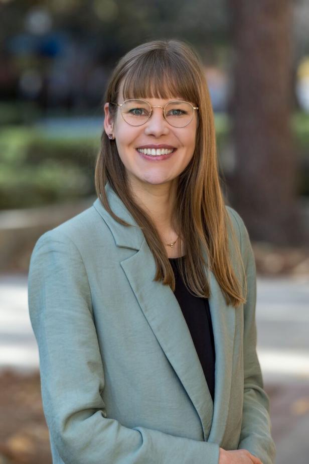 Eine lächelnde Frau mit Brille und Blazer steht im Freien.