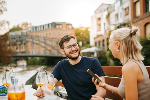 Ein junger Mann mit Brille und eine blonde Frau sitzen auf einem Boot und genießen Aperol Spritz. Sie fahren über einen Kanal in einer Stadt.
