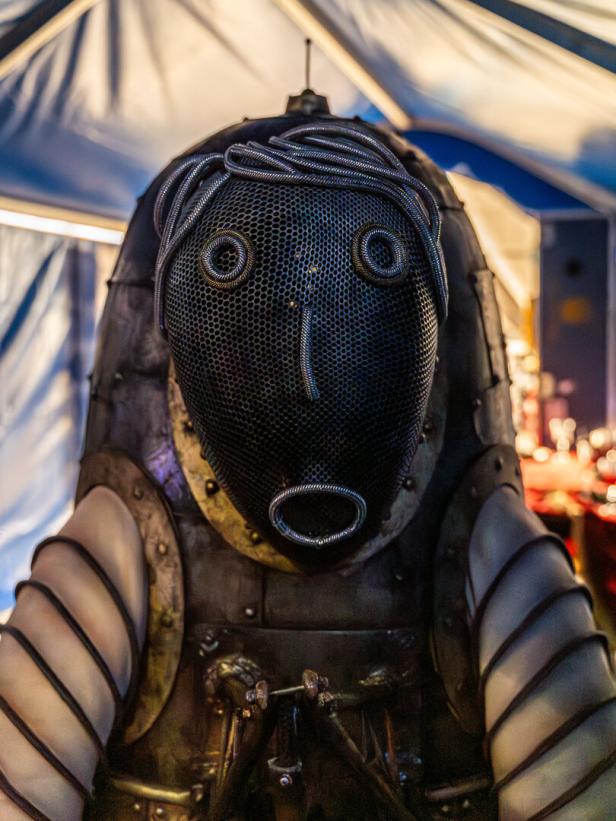 Eine skulpturale Figur mit einer Maske aus Metallgeflecht und drahtartigen Haaren.