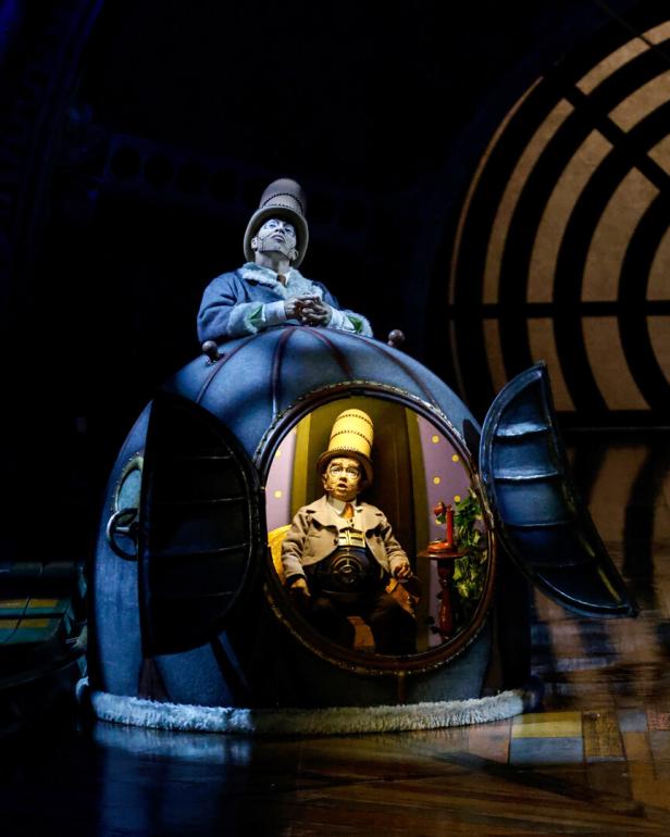 Zwei Figuren in Hüten sitzen in einer Art U-Boot auf einer Theaterbühne.