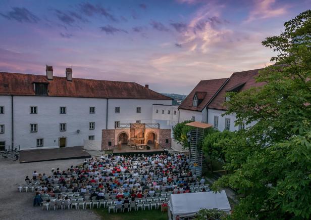 Der Hof einer Burg, in dem Dutzende Menschen in Reihen auf Stühlen vor einer Bühne sitzen. Die Sonne geht im Hintergrund unter.