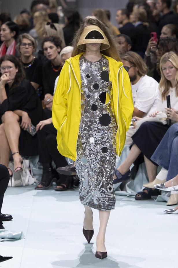 Ein Model auf dem Laufsteg trägt ein Paillettenkleid, eine gelbe Jacke und einen Lampenschirm-Hut.