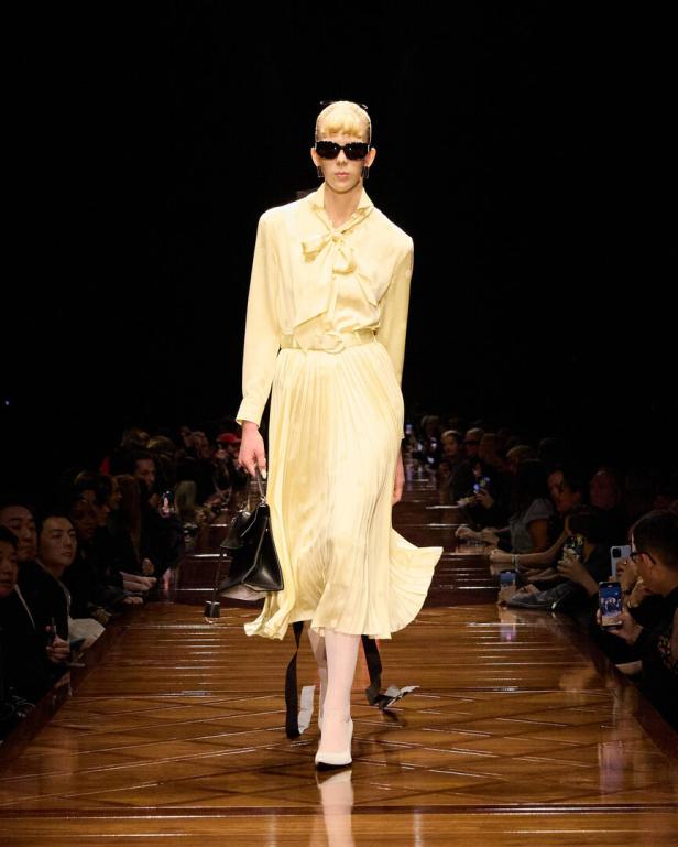 Ein Model in einem gelben Kleid mit Schleife läuft über einen Laufsteg.
