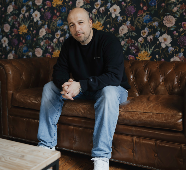 Ein Mann mit Glatze sitzt auf einem braunen Ledersofa vor einer floralen Tapete.