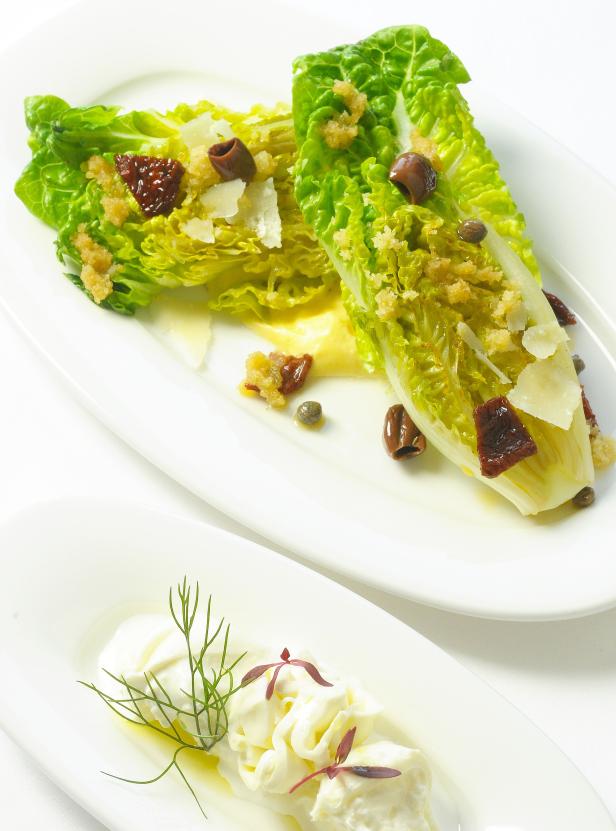 Zwei Römersalatherzen mit Parmesan, Oliven, Kapern und getrockneten Tomaten auf einem Teller.