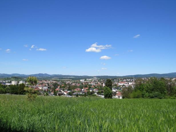 Grüne Wiese im Vordergrund, dahinter eine Kleinstadt mit Häusern und Bäumen, im Hintergrund Hügel unter blauem Himmel mit wenigen Wolken.