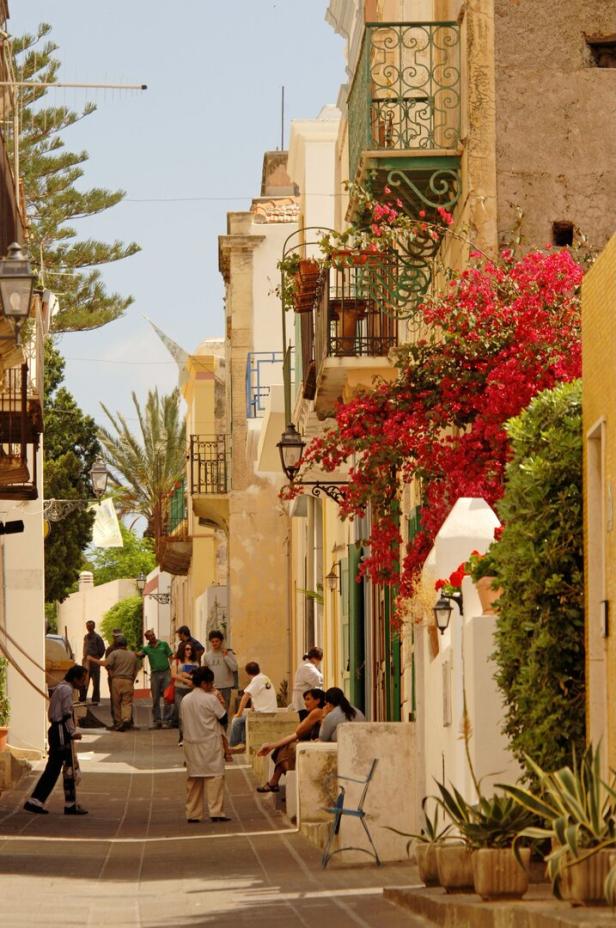 Eine belebte Gasse mit Menschen und blühenden Pflanzen in einer mediterranen Stadt.