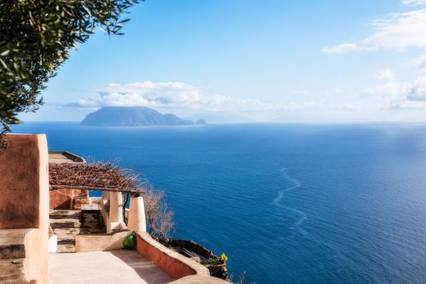 Blick von einer Terrasse auf das tiefblaue Meer mit einer Insel am Horizont.