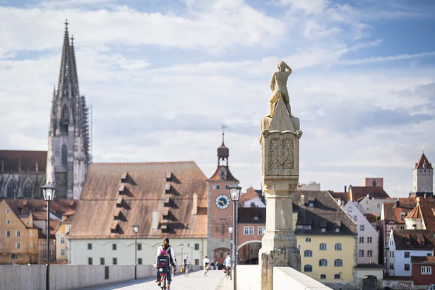 Ein Mann auf einem Fahrrad fährt über eine Brücke mit städtischer Kulisse und Dom im Hintergrund.