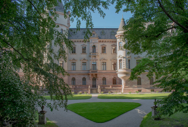 Großes Schloss mit grünem Rasen und Bäumen im Vordergrund, umgeben von einer malerischen Landschaft.