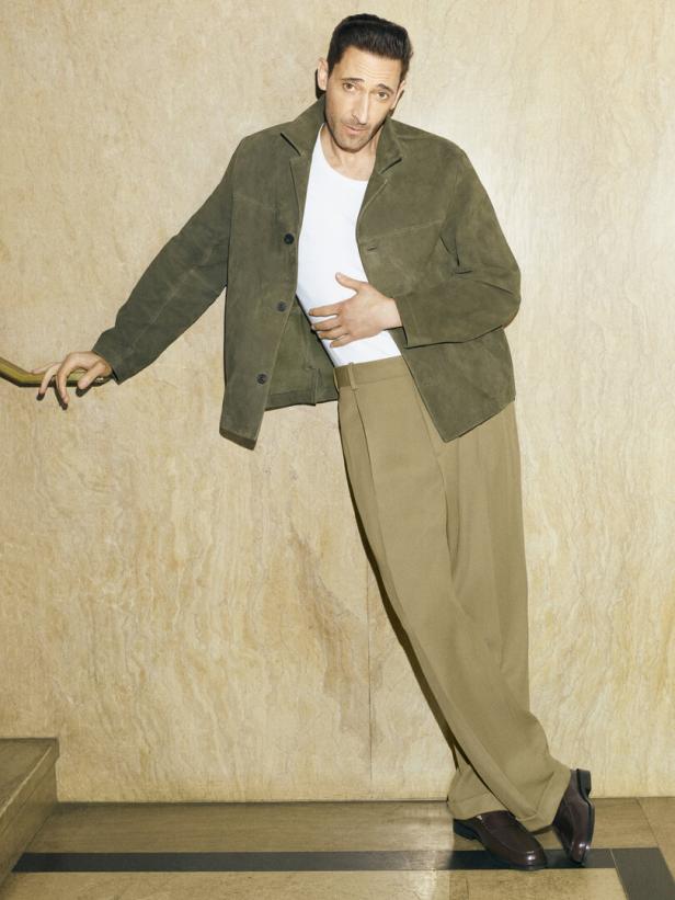 Adrien Brody posiert lässig an einer Wand mit Hand auf dem Bauch.