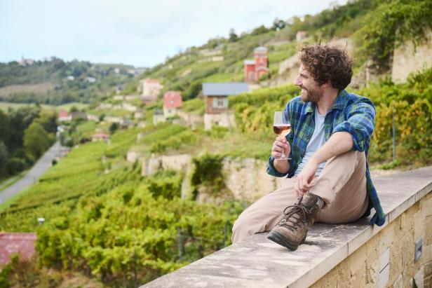 Ein Mann sitzt mit einem Weinglas auf einer Mauer und schaut auf Weinberge.