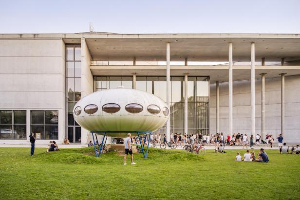 Ein modernes Museum von außen, mit einer Wiese davor, auf der Menschen sitzen.
